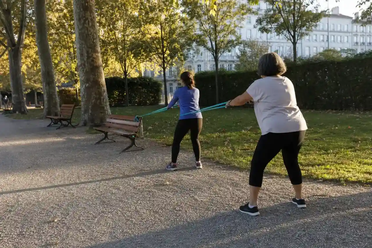 Deux femmes d’âges différents en séance de renforcement à l’élastique sur un banc de parc urbain lyonnais, lumière rasante de fin de journée