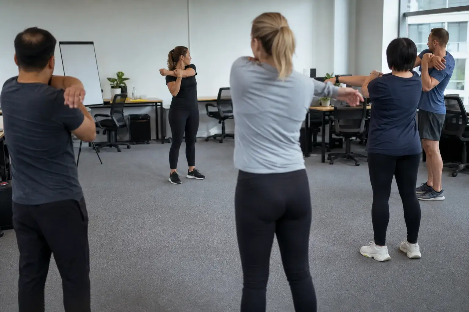 Cinq collègues en tenue de ville sportive en séance de mobilité d’épaules dans un plateau de bureaux dégagé, bureaux repoussés contre le mur