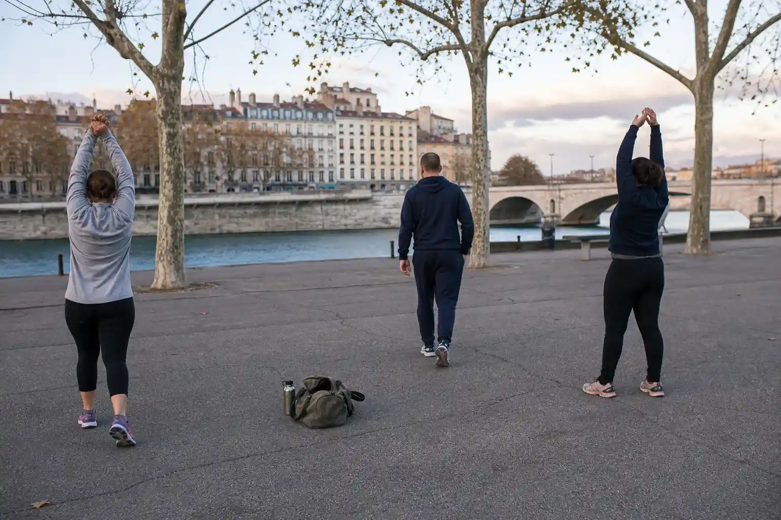 Coach sportif et deux adultes en fin d’échauffement sur un terrain public des berges du Rhône à Lyon, en fin d’après-midi