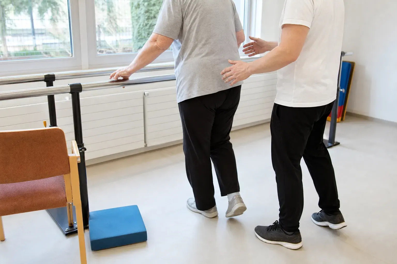 Travail d’équilibre d’une personne âgée à la barre parallèle basse, encadrante à côté, mains prêtes mais sans contact