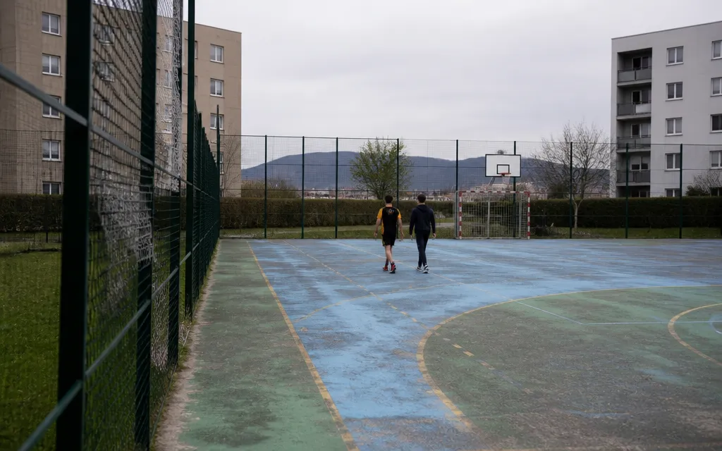 City-stade en plein air au pied d’immeubles bas de deuxième couronne, revêtement usé et grillage pare-ballons